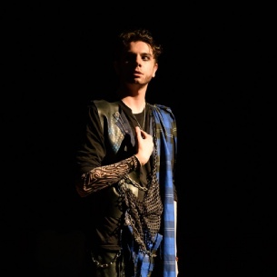 A person stands under dramatic lighting against a black background, wearing a costume with black and blue checked fabric and a tattoo-style sleeve, looking slightly upward.