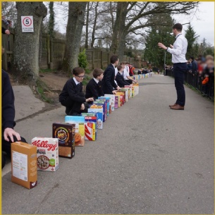 A group of students in uniforms kneel in a line outside, each with a cereal box in front of them, while an adult stands facing them and gestures with their hands. Trees and a fence are visible in the background.
