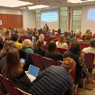 A large group of people sits in rows of chairs facing a speaker and presentation screen in a conference room. Most attendees appear to be women, and some are using laptops or taking notes.