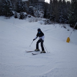A person wearing ski gear, including a helmet and goggles, skis down a snowy slope surrounded by trees, with a ski lift and snow-covered landscape in the background.