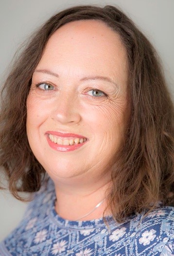 A woman with shoulder-length brown hair and blue eyes, smiling at the camera. She is wearing a blue patterned top and has a light background.