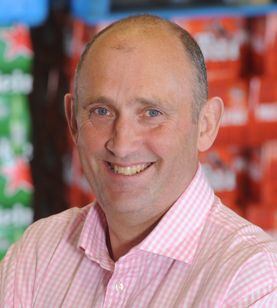 A middle-aged man with short hair smiles at the camera, wearing a pink and white chequered shirt. The background is blurred, showing stacked boxes in green and red colours.