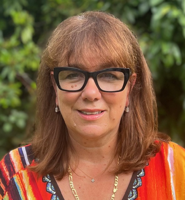 A woman with shoulder-length brown hair and black glasses smiles outdoors. She is wearing a colourful top with red, orange, and black stripes, a gold necklace, and earrings. Green foliage is blurred in the background.