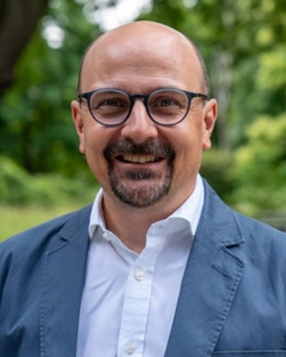 A smiling man with glasses, a goatee, and a bald head wearing a white shirt and a blue blazer, standing outdoors with green trees in the background.