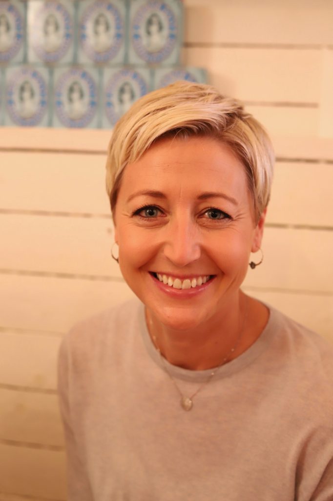 A smiling woman with short blonde hair and hoop earrings sits in front of a white wooden wall with a row of patterned tiles in the background. She is wearing a light-coloured top and a delicate necklace.