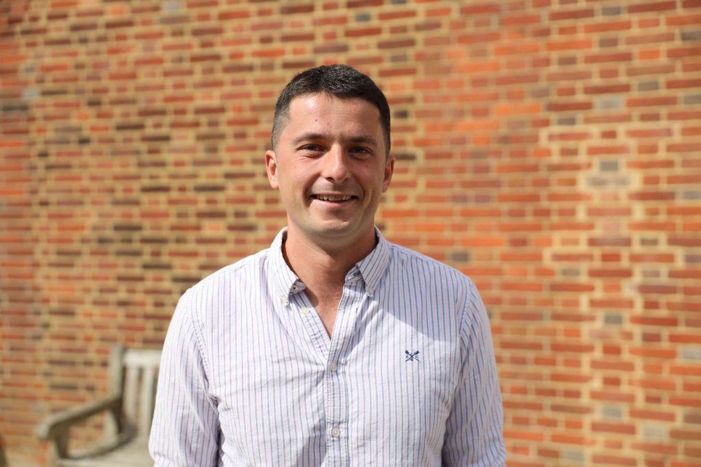 A man in a light-coloured, striped shirt stands outdoors in front of a sunlit brick wall, smiling at the camera. A blurred wooden bench is visible in the background.