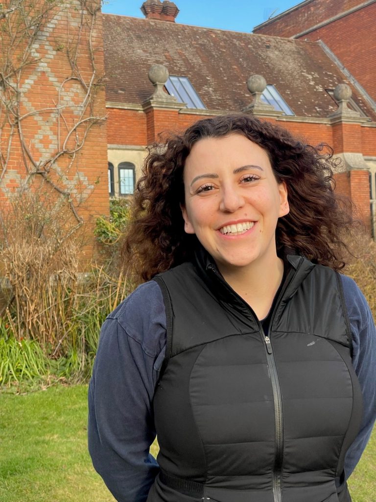A woman with curly brown hair wearing a black waistcoat and navy jumper smiles whilst standing outside in front of a brick building with large windows and a garden.