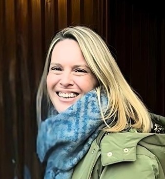 A woman with blonde hair smiles warmly, wearing a green jacket and a large blue patterned scarf, standing in front of a wooden background.