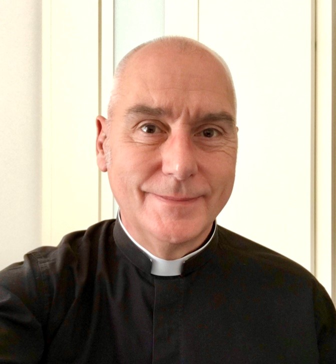 A middle-aged man with short, fair hair wearing a black clerical shirt with a white collar, smiling at the camera whilst standing indoors in front of light-coloured panels.