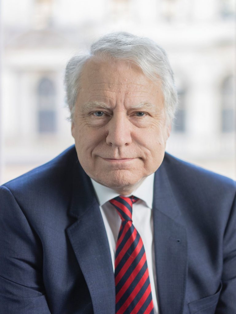An older man with grey hair is wearing a dark blue suit, white shirt, and red-and-blue striped tie. He is looking directly at the camera with a neutral expression, sitting in front of a blurred background.