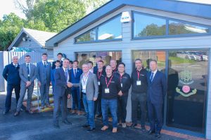A group of people, including men in suits and polo shirts, stand smiling outside the modern glass doors of the More House School St Matthew's building on a sunny day. Several men at the front shake hands.
