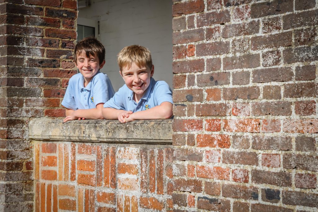 Two young boys in light blue polo shirts lean on a stone window ledge, smiling, as sunlight illuminates their faces—capturing a moment of joy amidst old brick walls, reminiscent of friendships formed during the EHCP process.