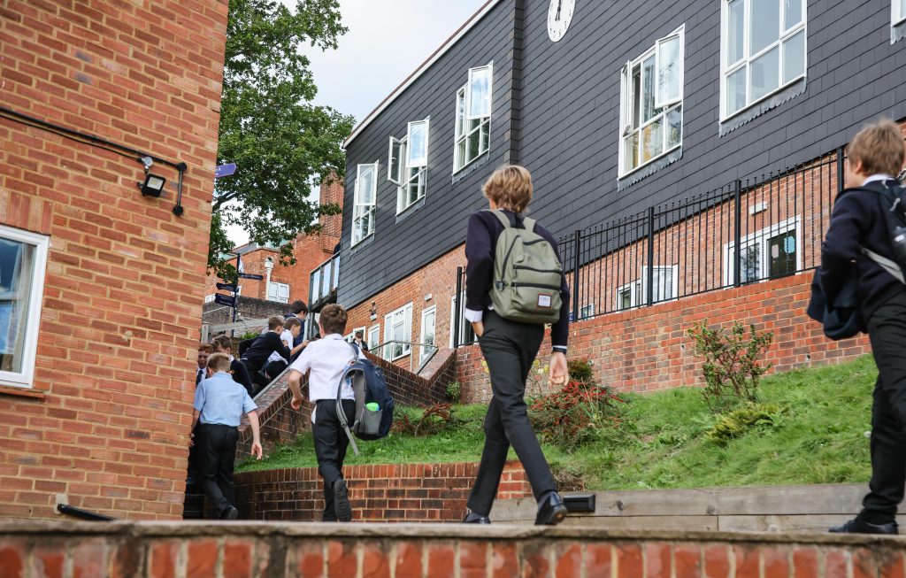 Several schoolchildren wearing rucksacks walk along a path between brick buildings, heading towards an entrance—some possibly beginning their EHCP process on the vibrant school campus.