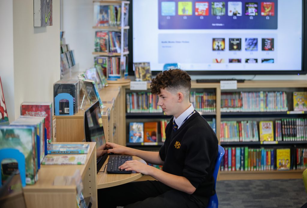 A student in a school uniform uses a laptop at a desk in the More House School Campus library, surrounded by bookshelves and a large screen displaying digital content in the background.