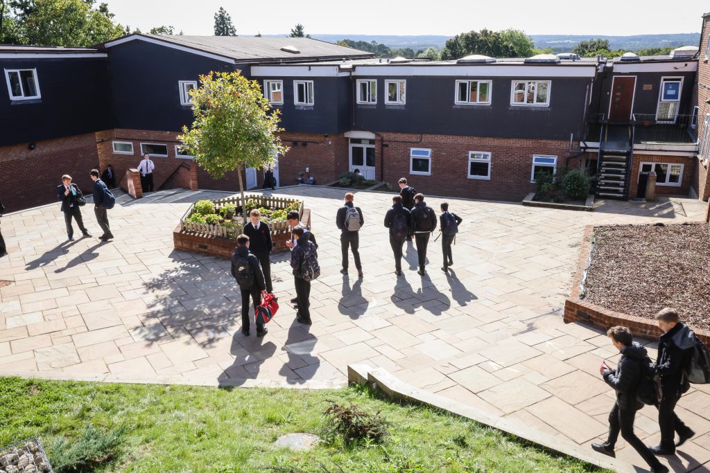A group of students in school uniforms stand and talk in a paved courtyard outside a two-storey school building on a sunny day, while some discuss the EHCP process with their peers. Some students are alone, whilst others are in small groups.