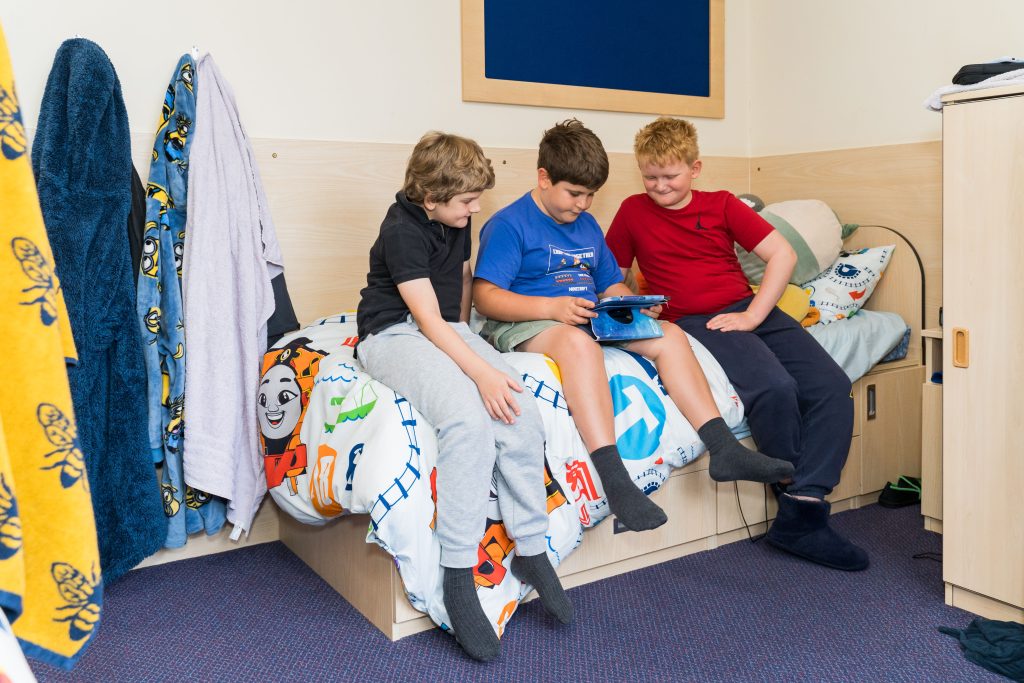 Three boys sit together on a bed in a cosy room, focused on a tablet device. The bed has colourful cartoon bedding, and the room has wooden furniture and a blue carpet, creating a cheerful, relaxed atmosphere.