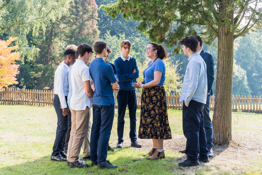 A female member of staff stands outside in the More House School Sixth Form back garden, speaking to a group of pupils who are gathered round her in under a tree.