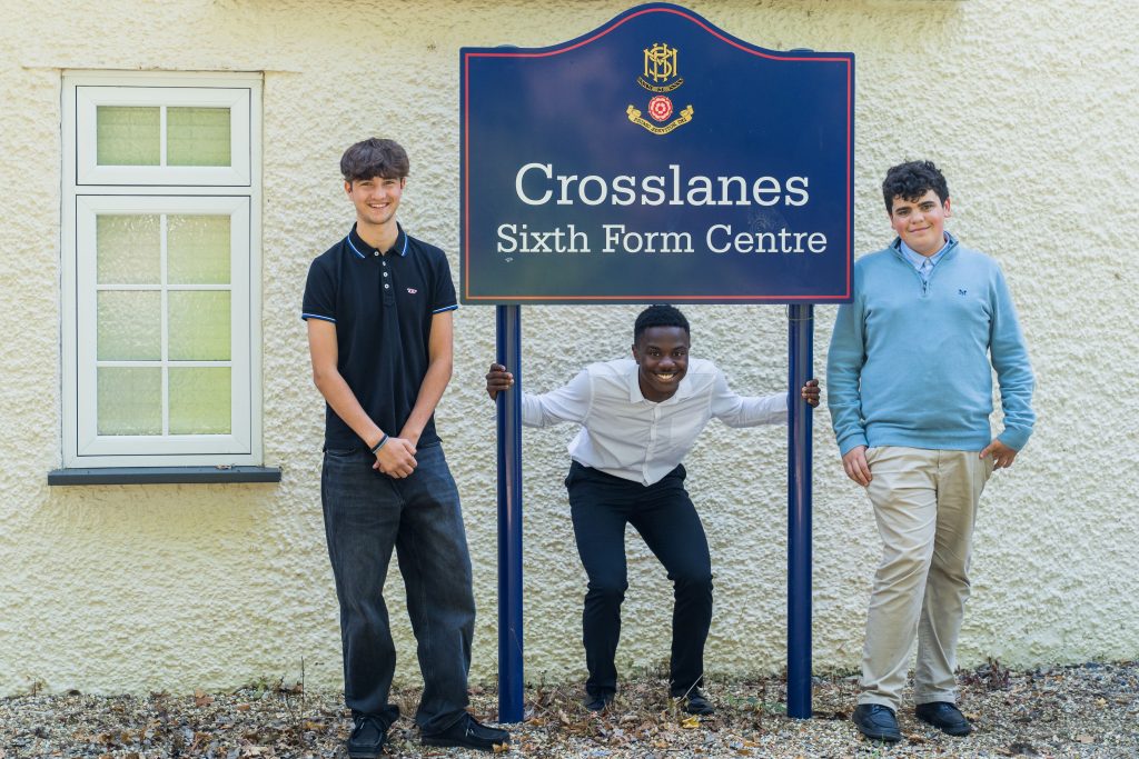Three teenage boys pose by a sign that reads “Crosslanes Sixth Form Centre” outside a cream-coloured building on the More House School Campus. One boy crouches and holds the sign while the other two stand on either side, smiling.
