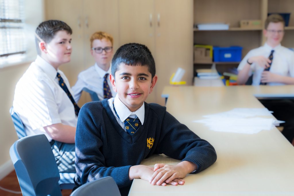 Four schoolboys in uniform sit at desks in a classroom. Using a School Specialist Approach, one boy at the front smiles at the camera while three others are seated behind him, two looking towards the camera and one looking away.
