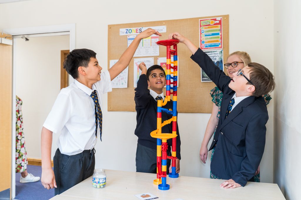 Three boys in school uniforms excitedly build a tall marble run with colourful pieces on a table, while a woman watches and smiles in a classroom setting on the More House School campus.