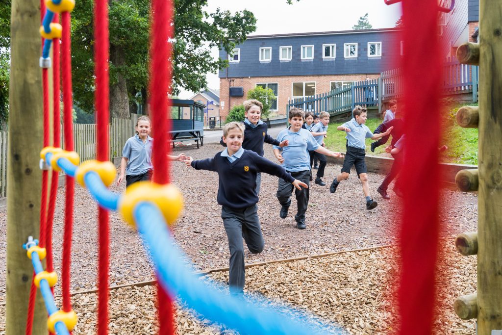 A group of children in school uniforms run and play outside on a playground, seen through colourful climbing ropes, reflecting a School Specialist Approach with the school building and trees in the background.