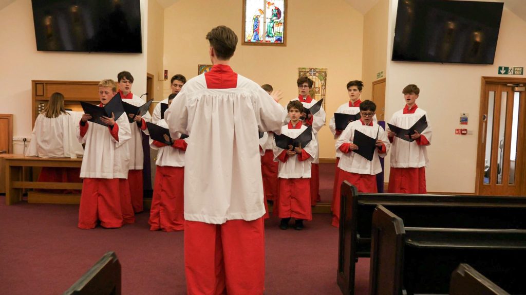 A choir of boys and young men in red and white robes sings in the school's Chapel, led by a teacher conducting them. The chapel has wooden pews, stained glass, and large screens on the walls.