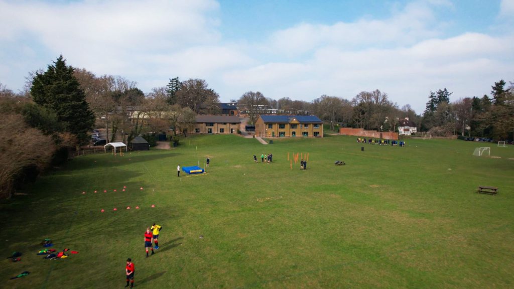 A large grassy field on the More House School campus, with pupils engaged in outdoor sports. Colourful markers and sports equipment scattered about, and a few buildings and trees in the background under a partly cloudy sky.