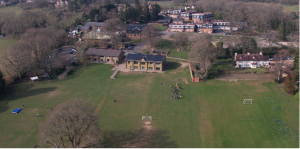 Aerial view of a grassy field with football goals, groups of people gathered near the centre, with several buildings surrounded by trees in the background.