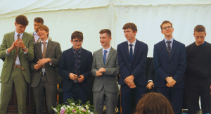 Seven young men stand in a row dressed in suits and ties, some smiling and clapping, under a white marquee. A bouquet of flowers sits in front of them, with a few people seated in the foreground.