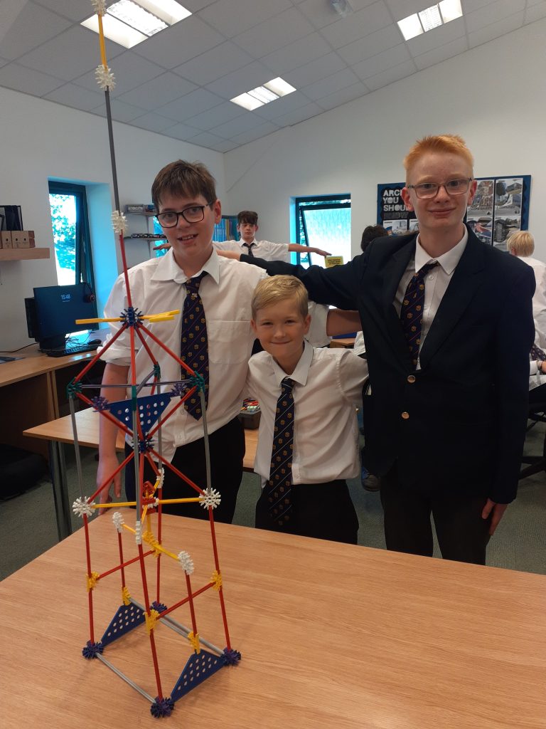 Three boys in school uniforms stand smiling behind a tall structure made of colourful plastic rods and connectors, placed on a wooden table in a classroom.