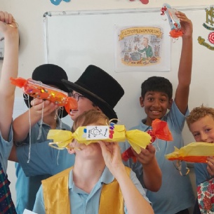 Small group of smiling children in school uniforms hold colourful homemade crafts, showcasing their creativity. One boy wears a black top hat, another a yellow waistcoat. They stand in a classroom focused on dyslexia and literacy support.