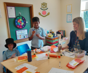 Three students and a teacher sit around a classroom table covered with papers, glue sticks, and craft supplies. Their smiles reflect the joy of creative learning in a space focused on dyslexia and literacy support.