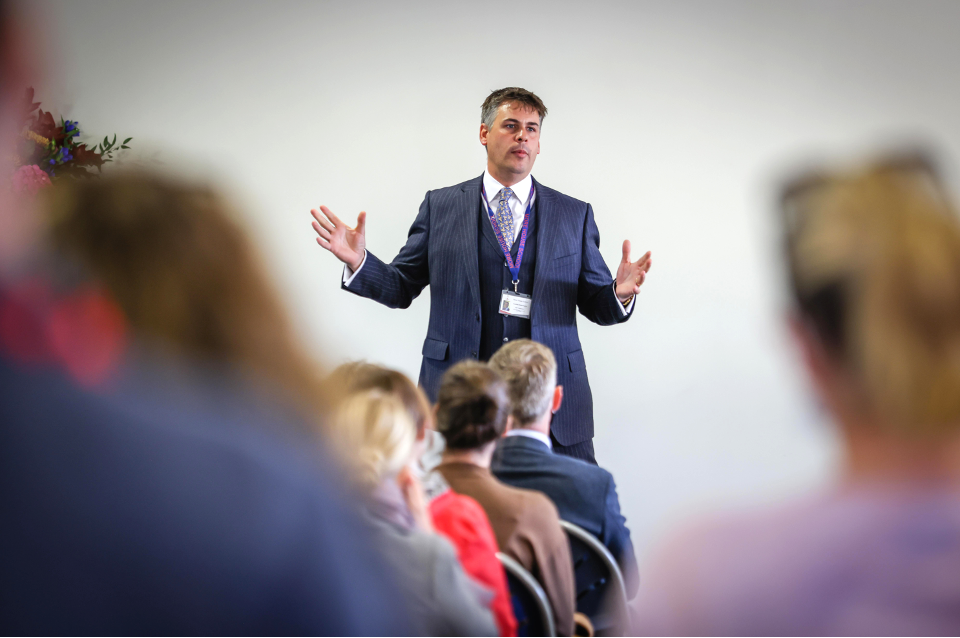 A man in a suit speaks to an audience, gesturing with his hands. The focus is on the confident speaker, reflecting his professionalism. The blurred audience highlights his commanding presence.