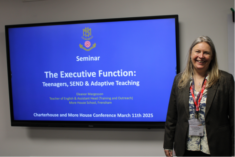 A woman in professional attire stands smiling beside a large screen displaying a seminar titled The Executive Function: Teenagers, SEND & Adaptive Teaching, highlighting consultancy and support at the Charterhouse and More House Conference, 11 March 2025.