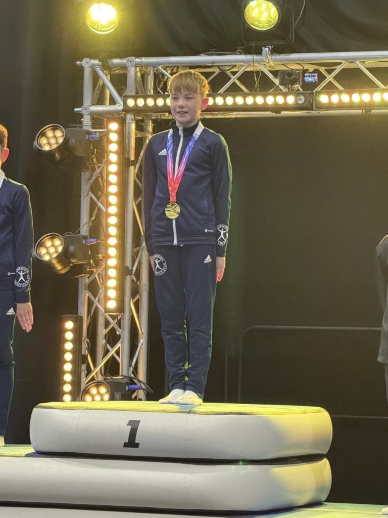 A boy wearing a tracksuit and gold medal stands on a podium marked 1, indicating first place.