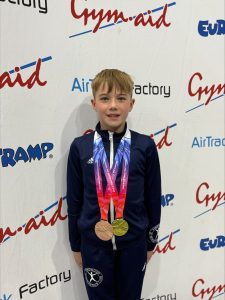 A young boy in a navy tracksuit stands smiling with two medals around his neck in front of a backdrop with Gym-Aid, AirTrack Factory, and Eurotramp logos.