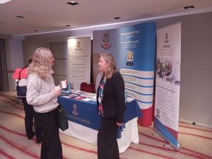Two women talk in front of a More House School information stand at an indoor event. The table has leaflets and banners about the school’s outreach, training, and inclusion services.