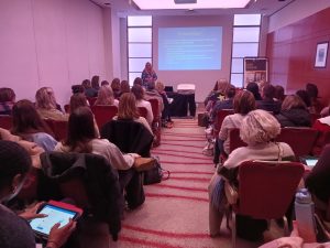 A woman stands at the front of a conference room, giving a presentation to an audience seated in rows. A projector displays slides on a screen behind her, and attendees look ahead or take notes on devices.