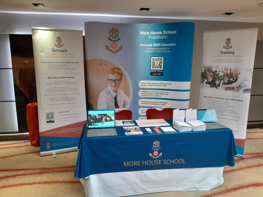 A promotional table for More House School is set up with brochures, a laptop, and display banners about their SEND training, outreach, and education services. The table is covered with a branded tablecloth.