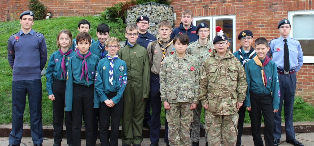 A group of sixteen young people in various scouting and cadet uniforms stand outdoors in front of a brick building and grassy hill, posing for a group photo.