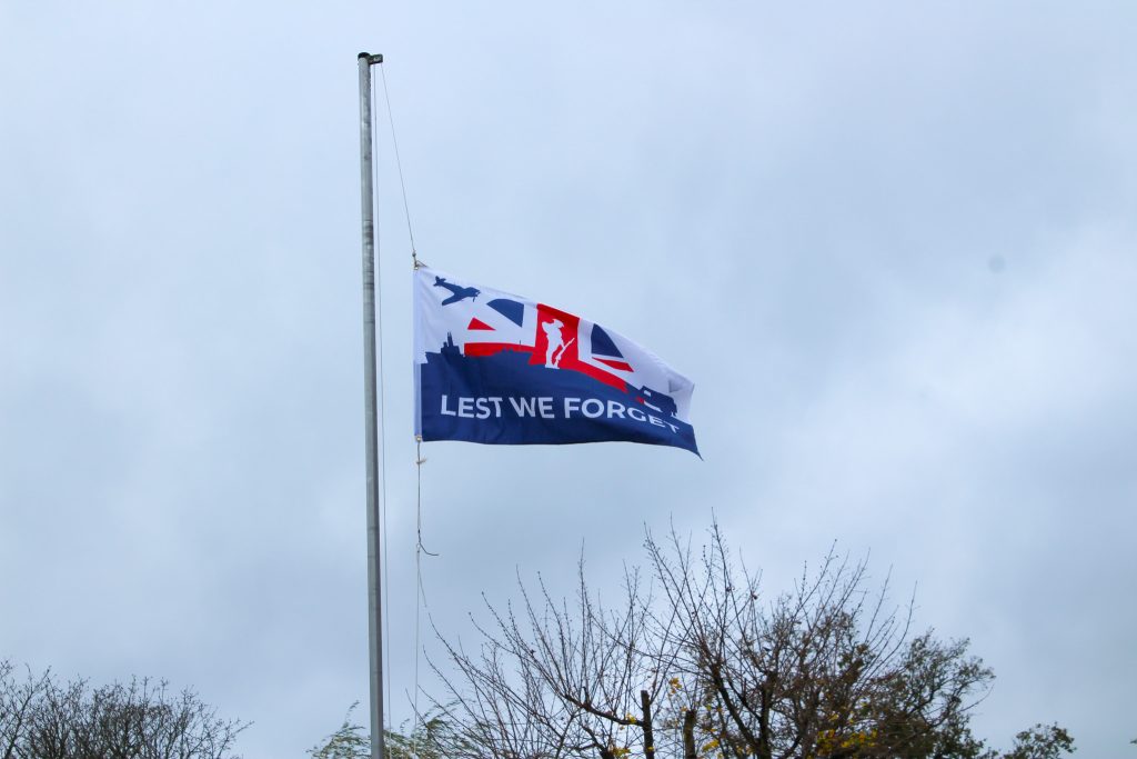 A flag with the words LEST WE FORGET flies on a flagpole against a cloudy sky, with silhouetted trees and branches visible at the bottom of the image.