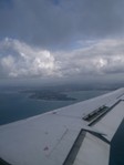 View from an aeroplane window showing the wing, clouds in the sky, and a coastline with land and water below.
