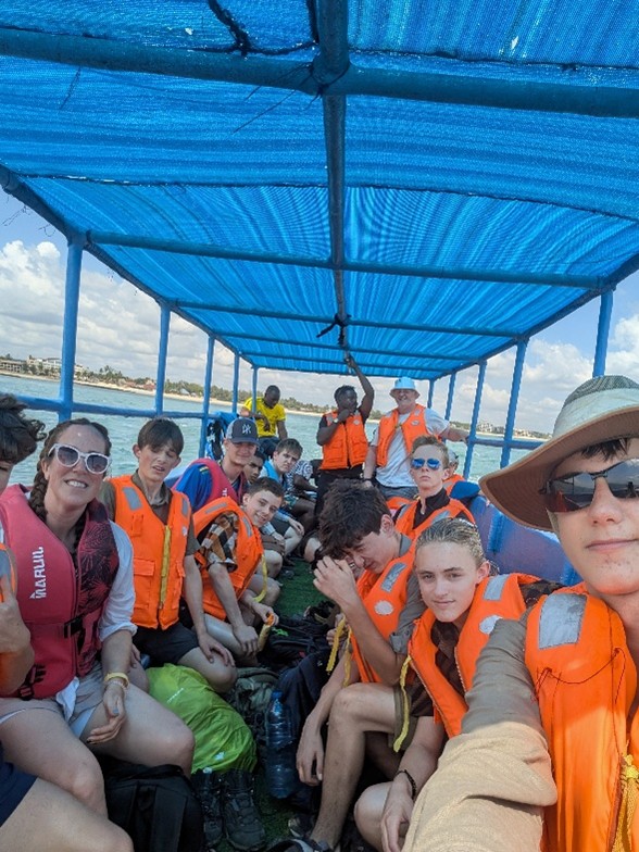 A group of people wearing orange life jackets sit closely together on a covered boat under a blue tarpaulin roof, with water and sky visible in the background. Some smile at the camera whilst others look relaxed or thoughtful.