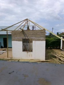 A person stands on the wooden roof frame of a partially constructed house with white walls and unfinished masonry, surrounded by scattered building materials and clouds in the sky above.