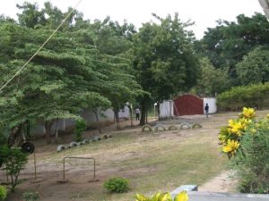 A grassy garden with scattered trees, a row of tyres partially buried in the ground, metal playground equipment, and a large red gate in the background. Two people are standing near the gate and foliage surrounds the area.