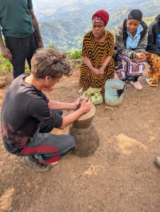 A young man kneels and carves a wooden object outdoors, while three women and another man watch. The women sit on the ground with bags and containers, overlooking a scenic, mountainous landscape in the background.
