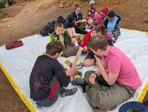 A group of people sit cross-legged on a white tarpaulin outdoors, sharing a meal from communal plates placed in the centre. Some wear casual clothes while others have colourful headwraps. The ground is earthy and rocky.