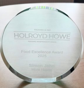 A glass trophy engraved with Holroyd Howe Food Excellence Award 2025 and Simon John, More House. The company slogan Feeding Independent Minds is also visible above the award text.
