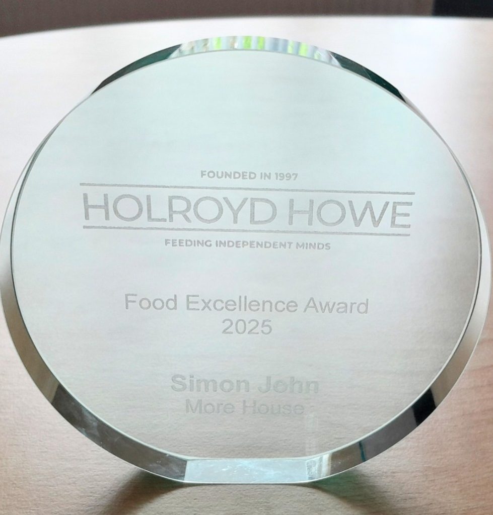 A glass trophy engraved with Holroyd Howe Food Excellence Award 2025 and Simon John, More House. The company slogan Feeding Independent Minds is also visible above the award text.
