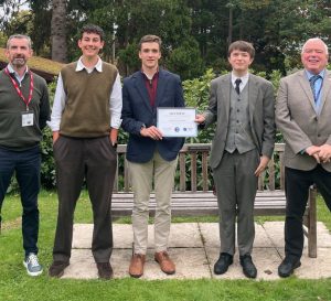 Five men stand outdoors on grass in front of a wooden bench. The central man holds a certificate. All are dressed smartly in blazers or suits, and they are smiling, with trees and bushes in the background.